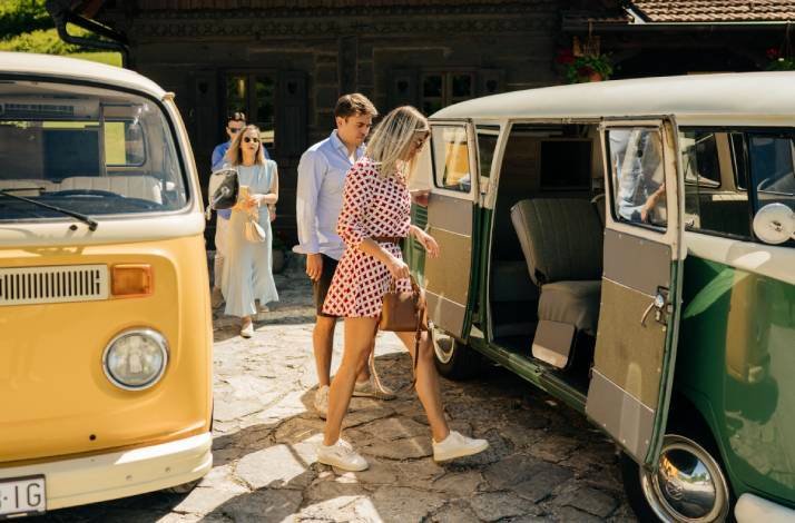 A group of people gets into a vintage van parked outside a rustic building.