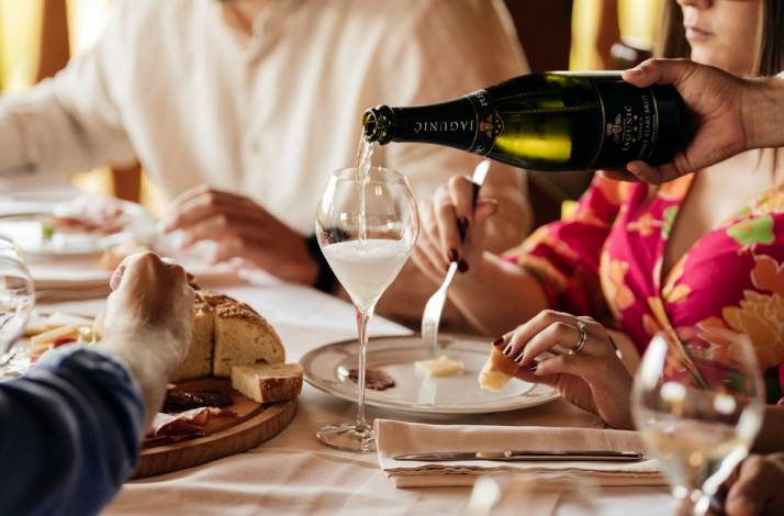A person pours sparkling wine into a glass at a table with bread and cheese.
