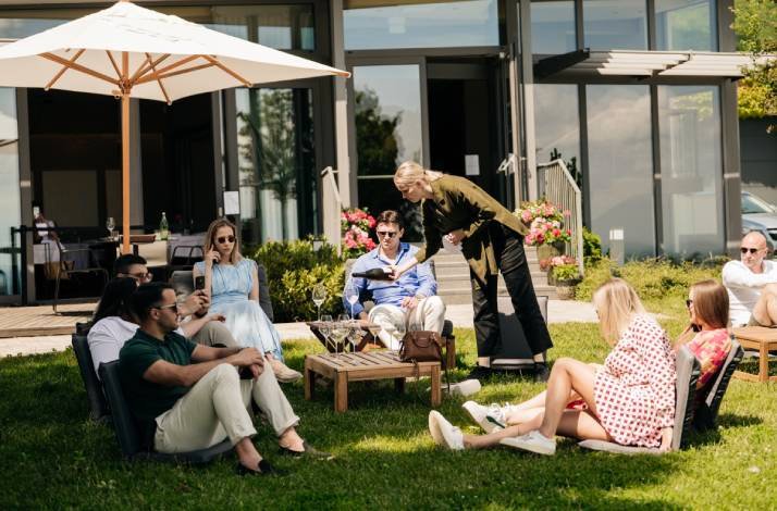 A group of people relaxes on lawn chairs under a parasol while being served drinks.