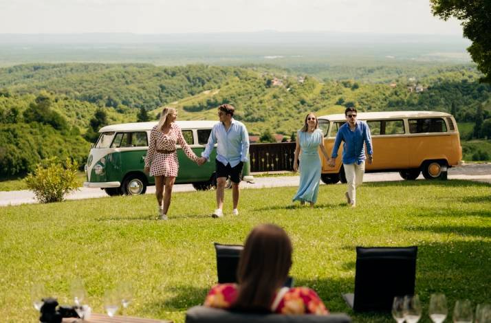 Four friends walk across a grassy hill with vintage vans and scenic views behind them.