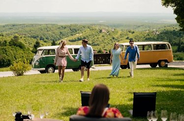 Four friends walk across a grassy hill with vintage vans and scenic views behind them.