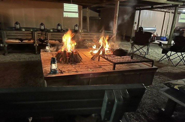 A campfire is burning in a rustic outdoor setting, surrounded by wooden benches, foldable seats and camping equipment at Kruger National Park