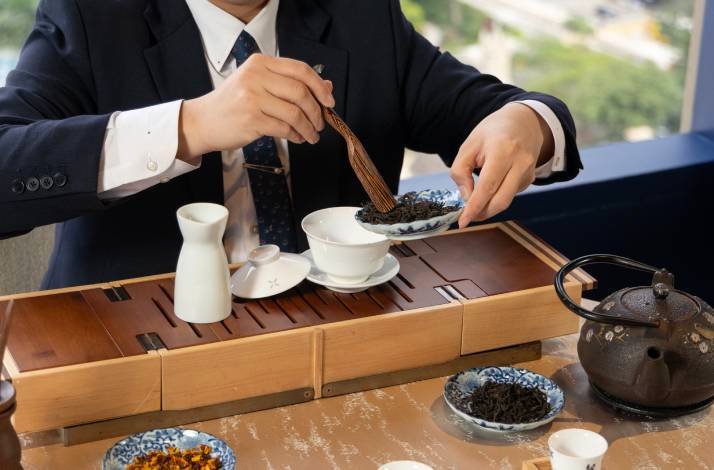 A man in a suit places loose tea leaves into a traditional Chinese gaiwan.