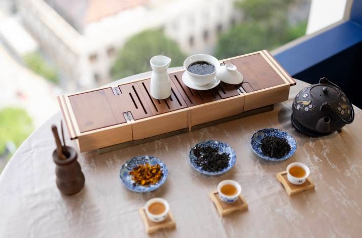 A tea setup with loose leaves, cups, and a gaiwan on a table near a window.