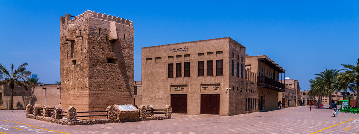Ancient mud-brick watchtower and restored heritage buildings in Old Dubai's Al Fahidi district under a clear blue sky.