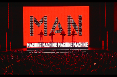 Kraftwerk performing live with red backdrops spelling MAN MACHINE in front of a seated audience.