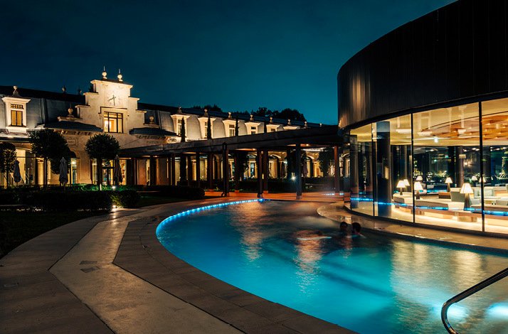 Outside pool surrounding a large lounge room with crystal windows at night.