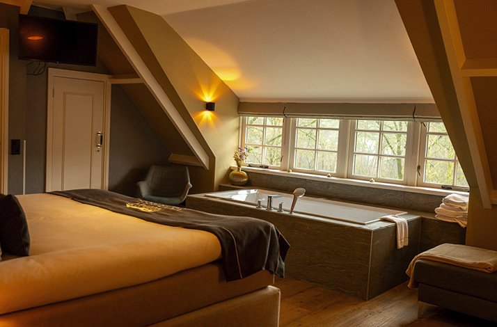 An interior photo of the Landgoed de Uitkijk  hotel room, equipped with a queen sized bed, facing a bathtub and several glass windows. Several towels are situated beside the bathtub.