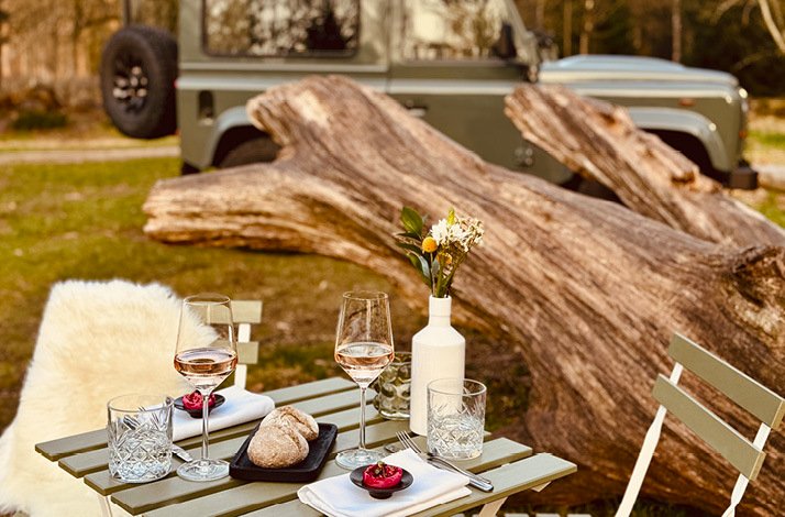 A photo of some plated foods, glasses of drinks, a flower vase and utensils served on an outdoor wooden table along with outdoor wooden chairs where one is covered with wool cover. The surrounding of the table is paired with a big wood log and a 4x4 jeep truck in an outdoor setting.