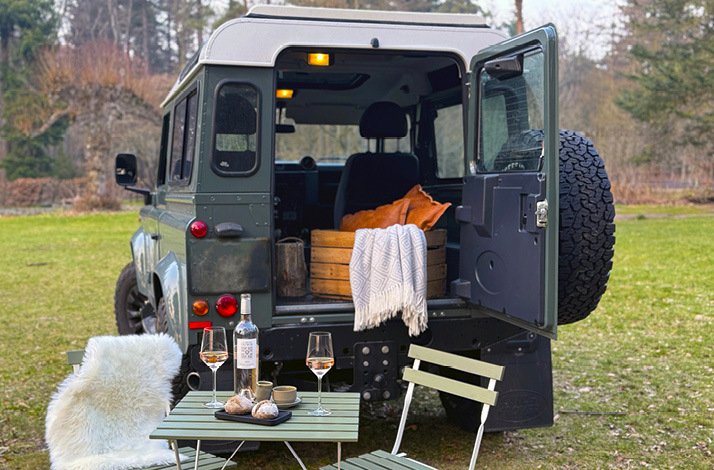 Photo of an open trunk 4x4 jeep truck filled with home products, nearby the truck is some plated foods and glasses of drinks served on a wooden outdoor table along with chairs covered with wool cover