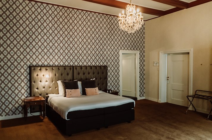 A bedroom shot at Chateauhotel de Havixhorst, which features a king-sized bed placed on the diamonds and floral patterned wallpaper wall, a chandelier, and a brown carpeted floor. The surrounding walls are a neutral color, doors are visible at each end, and a small table is at the bedside of the bed. 
