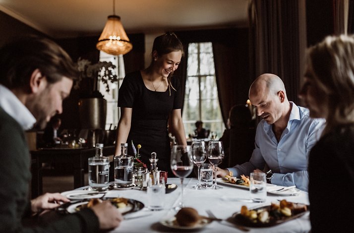 A group of people are seated around a table in Chateauhotel de Havixhorst's restaurant, enjoying a meal, and being served by a staff member. The scene is composed around a large, round table with dishes, glasses, and serving platters, and the guest's dress code suggests a formal or semi-formal dining setting.