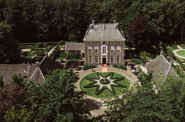 An aerial view of Chateauhotel de Havixhorst, featuring a circular flowerbed design garden situated in front of the hotel's entrance. The entrance is surrounded by various large estate with other gardens, and an orange carpet leads to the entrance of the hotel.