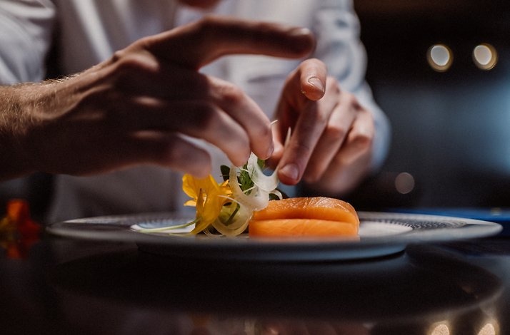 A close-up view of a chef's hands carefully arranging garnishes, which includes edible flowers in yellow and white along with other greens, on a plate of orange salmon slices.