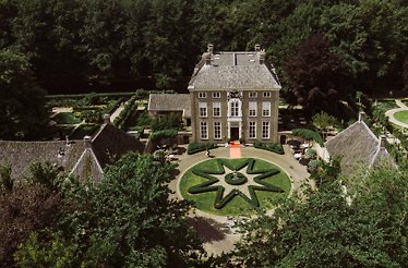 An aerial view of Chateauhotel de Havixhorst, featuring a circular flowerbed design garden situated in front of the hotel's entrance. The entrance is surrounded by various large estate with other gardens, and an orange carpet leads to the entrance of the hotel.