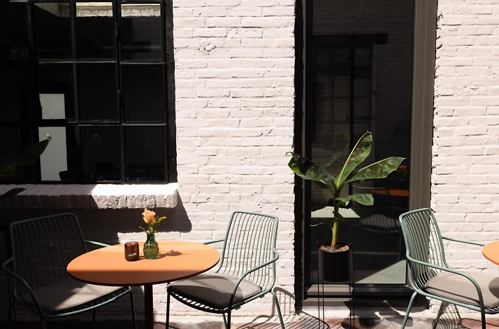 An outdoor patio area of Cousins Boutique Hotel, featuring a round orange table, complemented with green chairs, situated against white brick walls. A small banana plant pot is situated nearby the table, and a mini flower vase sits on the table. 