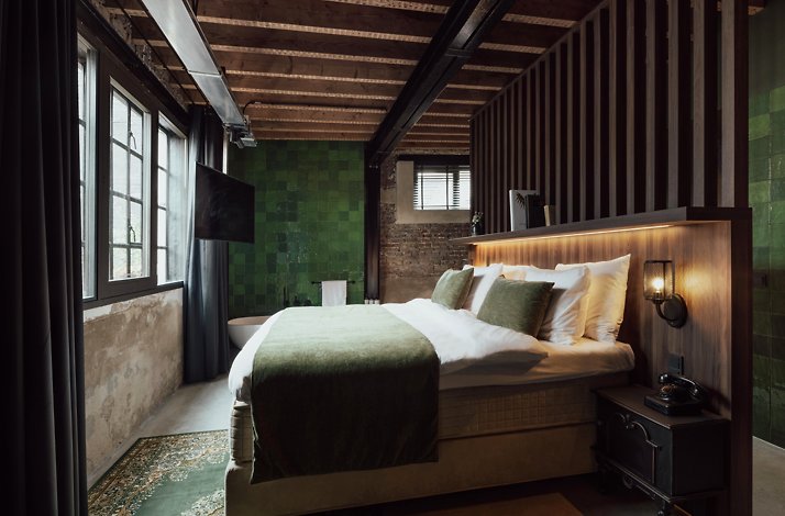 A bedroom interior of Cousins Boutique Hotel, featuring a king sized bed, complemented with a dark-green bedspread and pillows. The room's walls,  featured a dark wooden headboard and panels, incorporating an element of industrial design of brick walls, green tiles and exposed beige colors. The windows are placed in front, and a bathtub is placed to the right of the bed.