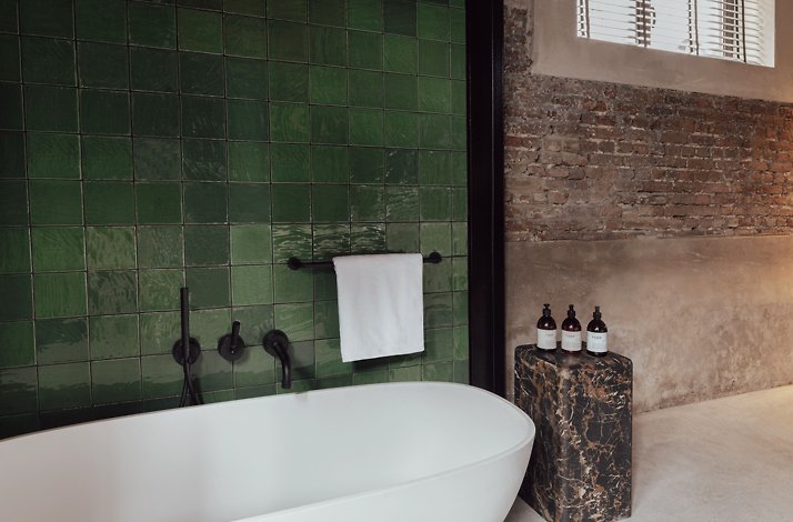 A close up shot of the interior of Cousins Boutique Hotel's bathroom, featuring a white bathtub, with a dark-colored metal towel rack mounted on the wall with towels, and a dark stone pedestal with several bottles, placed beside the tub. The walls are designed of partly squared green tiles, exposed brick and beige colored wall.