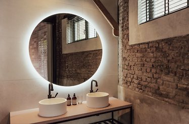 A close up shot of the interior of Cousins Boutique Hotel's bathroom, featuring a round vanity mirror with two rounded sinks. The bathroom walls are of partly exposed brick and beige in color, providing an industrial feel.