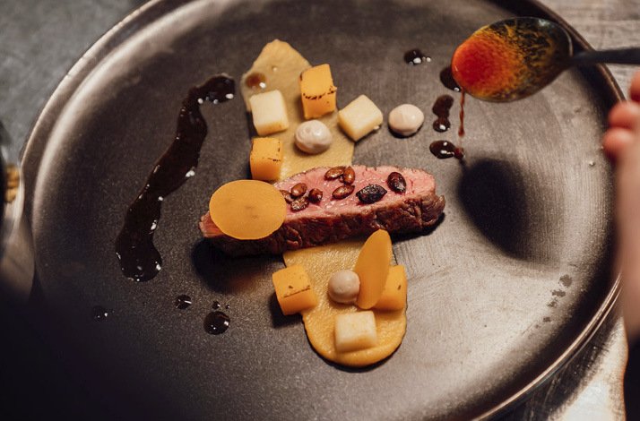 A closeup image of a plated dish offered by Broederenklooster Hotel, which includes of sliced meat, garnished with colorful vegetables and sauces, presented on a dark plate.