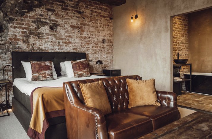 A photo of Broederenklooster Hotel's bedroom suite decorated in exposed brick walls, along with a king-sized bed and a dark brown leather sofa paired with light brown pillows placed to the front of the bed. 