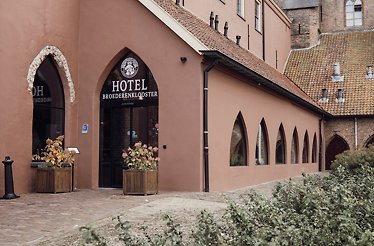 A shot of the exterior entrance of Broederenklooster Hotel in light pinkish color, where the entrance door is decorated with two flower plants in wooden pots beside it.