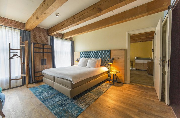 An interior of Van Rossum Woerden's hotel room, equipped with a king sized bed, where beside the bed includes a connecting room door, two side bed lights, a garment wooden cabinet and rack