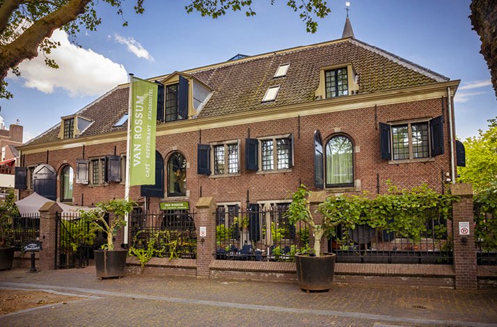 A photo of Van Rossum Woerden front building, with a yellow green banner beside the front gate, that says Van Rossum Cafe Restaurant Stadshotel
