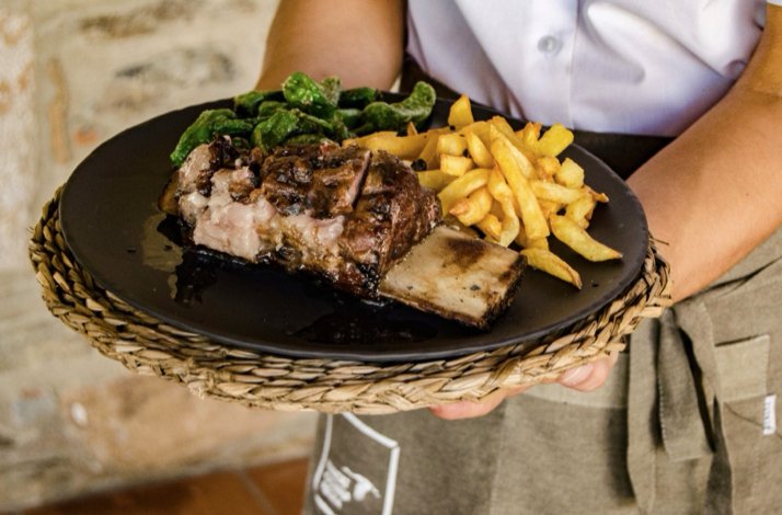 A server presents a dish with grilled beef ribs, fries, and green peppers on a black plate.