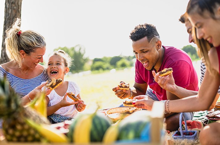 A family happily enjoying a picnic on a sunny day in the park, surrounded by trees and a blanket of food.