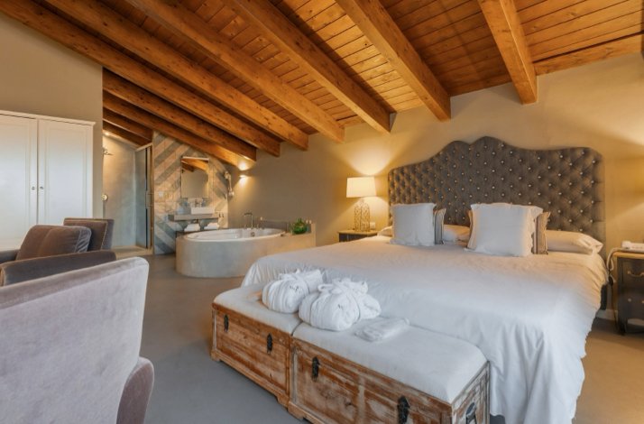 The Grand Suite Deluxe with a large bed, wooden beams, a seating area, and an open bathroom with a bathtub.