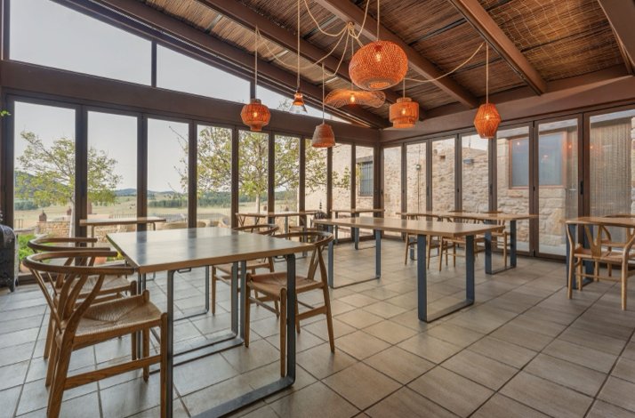 A dining area with wooden tables and chairs, large windows offering countryside views, and hanging woven light fixtures.