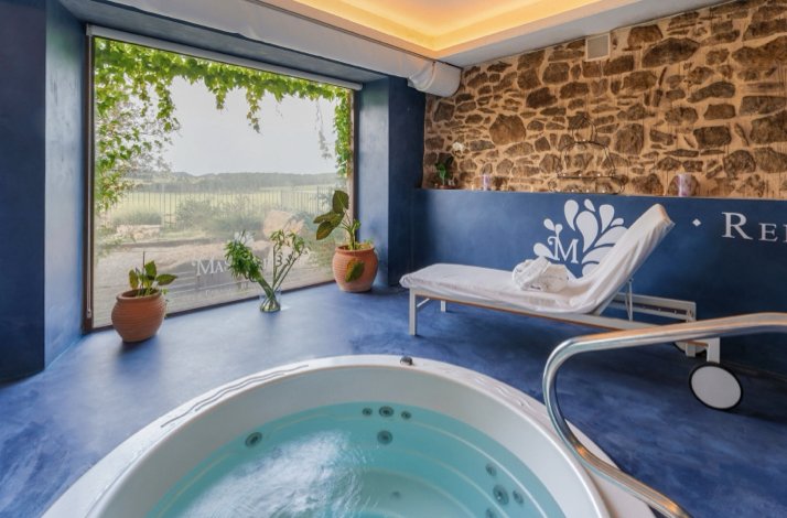 A private spa room for two with a whirlpool tub, lounge chair, stone walls, and a large window offering views of Teruel's natural landscape.
