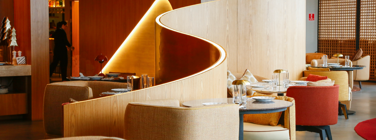 Taste of Priceless lounge interior at Guarulhos Airport, featuring set tables, warm-toned seating, and a sculptural wood partition.