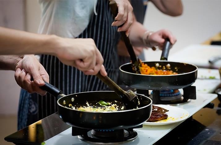 Two people are cooking in a kitchen of Studio Cooking Class, stirring food in frying pans on a stove.