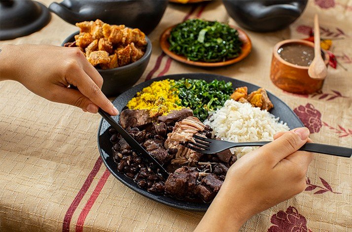 A person is seen cutting into a plate of Feijoada, a traditional Brazilian dish with black beans, rice, and pork.