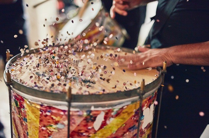 A close-up view captures people playing a drum, with colorful confetti flying around.