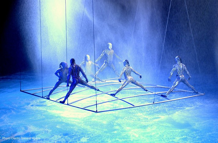 Artists in zebra-like suits perform intricate choreography on a suspended grid under water effects. Photo Credit: Tomasz Rossa