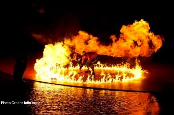 Performer surrounded by dynamic flames over water in a dramatic fiery display. Photo Credit: Julie Aucoin