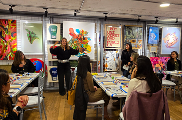 Art workshop with attendees seated, vibrant paintings, posters, and creative tools displayed around the room