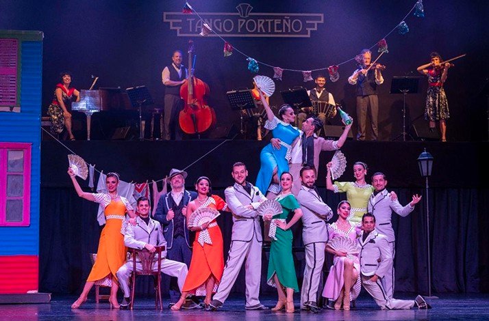 A lively tango scene with dancers in vibrant costumes and festive decor, backed by live musicians on stage of Tango Porteño Performing arts theater.
