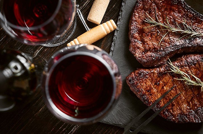 Grilled steaks with rosemary and glasses of red wine on a dark table.