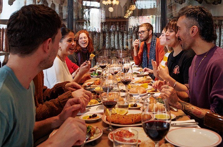 Group dining at a table with red wine, cold cuts, and bread in a charcuterie-filled room.