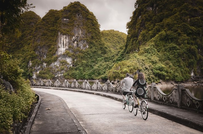 A couple is seen riding tandem bicycles on a road in Hạ Long Bay's Heritage Line surrounded by lush greenery and rocky hills.