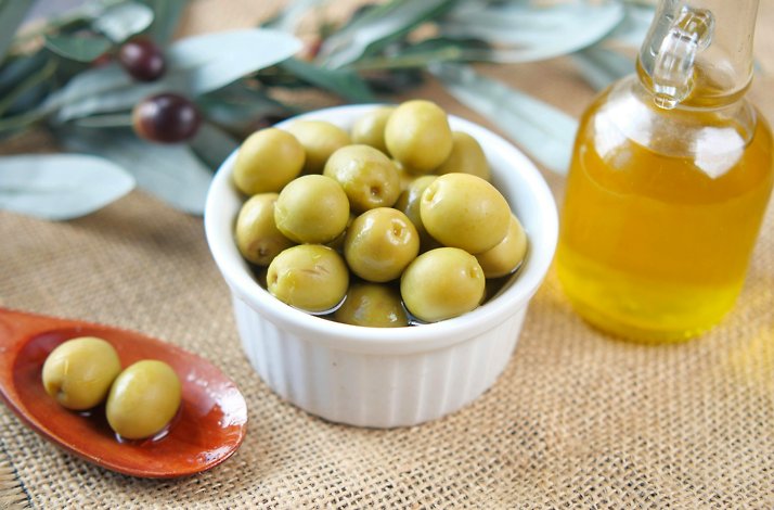Bowl of green olives with olive oil bottle, wooden spoon, and leafy background