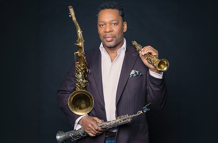 Jazz saxophonist Ravi Coltrane posing with multiple saxophones against a dark background.