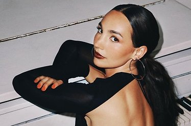 Emily Bear seated at a white piano, wearing a backless black outfit and hoop earrings, glancing over her shoulder.