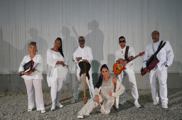 Promotional photo of percussionist Sheila E, labeled the Queen of Percussion, in front of her accompanying band.