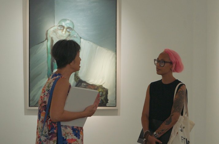 Angeline Gong and an art enthusiast discuss paintings with dark, emotive imagery displayed on minimalist white gallery walls