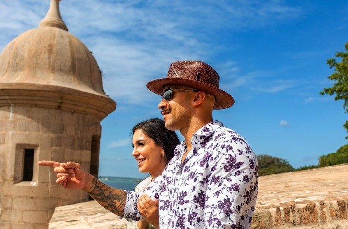 A couple stands at the Castillo San Felipe del Morro, pointing towards a distant vista under a clear blue sky.
